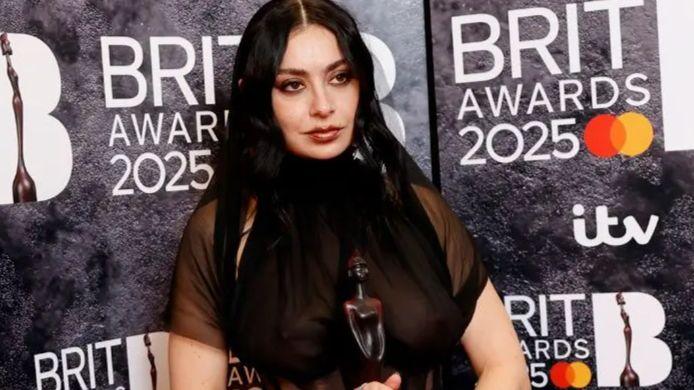 Charli XCX in her sheer dress holding a Brit Award in front of a Brits-branded backdrop