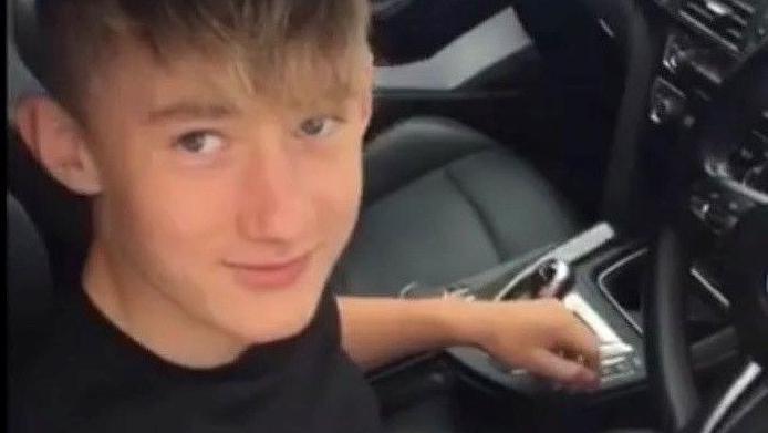 A boy wearing a black top sits in the driver seat of a car, posing for the camera.