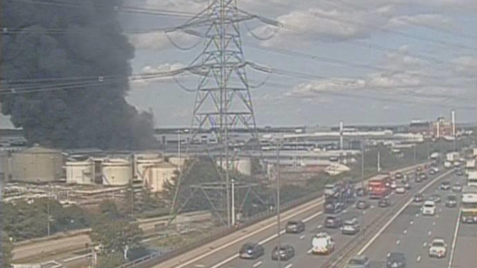 Traffic cam image of black clouds of smoke rising into the sky, near an oil terminal. Vehicles are passing along the motorway nearby