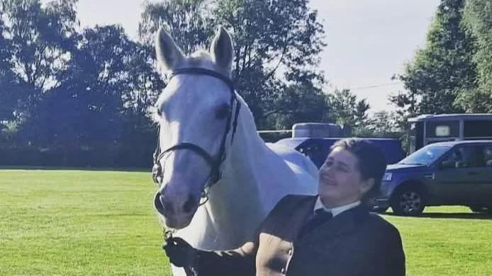 Poppy Bell holds Bess by the reins and stands next to her. Poppy wears an equestrian green blazer with a white shirt and tie. Bess is a white horse and has a black bridle on.