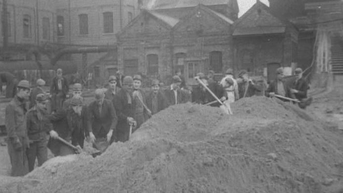 A black and white image of dozens of men filling up sand bags with shovels.