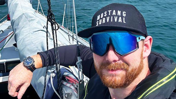 Kyle is on a boat and wears a navy blue long-sleeved top, blue sunglasses and a navy blue cap 