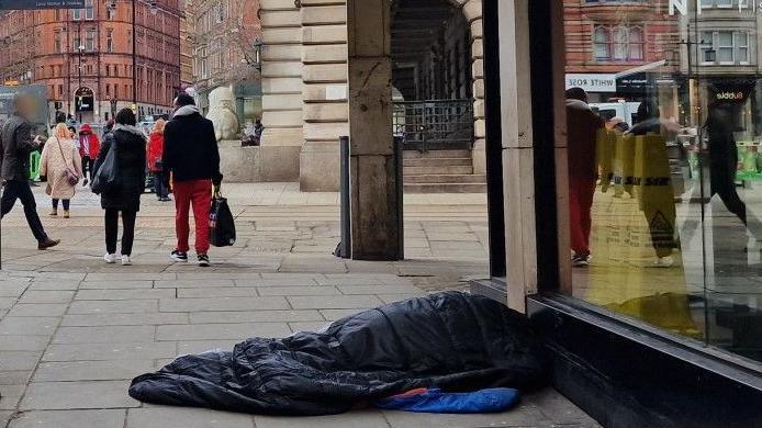 A pedestrianised road in Nottingham city centre showing the market place in the background. Shoppers pass a figure lying in a sleeping bag