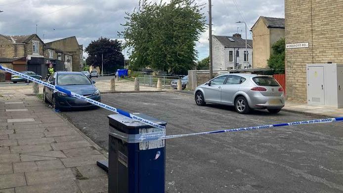 Police tape around a scene in a street. Two cars are pictured on the road, which comes to a dead end against a pavement. A police officer and houses can be seen in the distance.