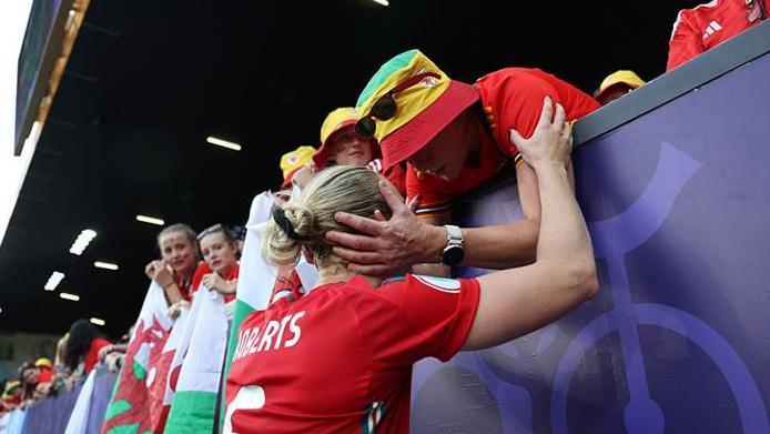 Rhiannon Roberts with her back to the camera. She is at the audience stand holding on to a woman's arm. The woman is holding on to Rhiannon's face and is consoling her while other fans to her left look on.