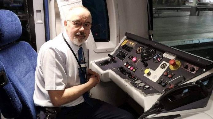 Don Coffey sat in the train driver cabin with a dashboard in front of him. He is wearing a white short-sleeved shirt and dark tie.