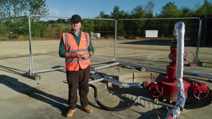 A man in an orange high-viz jacket stands beside a well with red and silver pipes and cylinders coming from it.