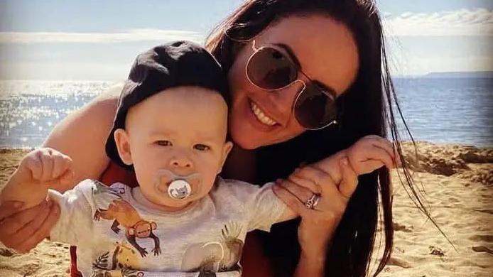 A young woman with dark hair holds up a baby boy in a sun hat and dummy on the beach.