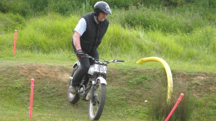 A one armed man rides a trail bike over a grassy hill marked by red posts. He wears a large black bike helmet