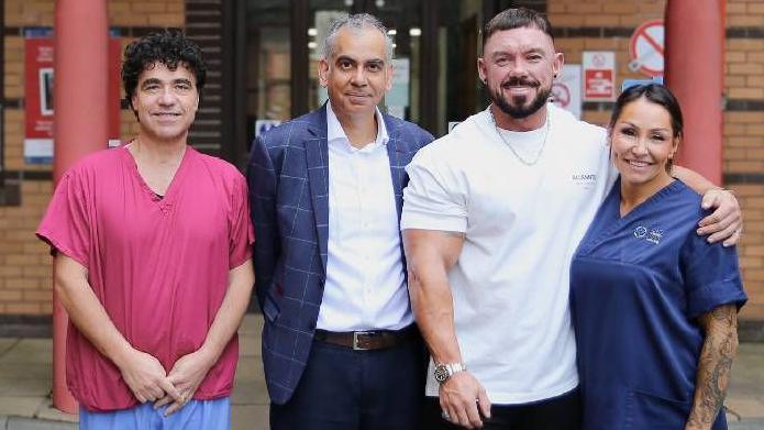 Neil Andrews, wearing a white T-shirt, with three other medical staff standing outside a hospital. Two - a man and woman - are wearing hospital scrubs, and another man is wearing a suit jacket and a white open shirt. 