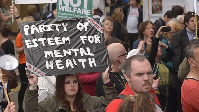 People demonstrating in the street in Manchester, England. One campaigner is holding up a placard that says "PARITY OF ESTEEM FOR MENTAL HEALTH".