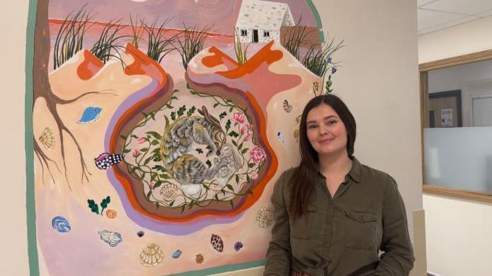 Ms Stuchfield stands next to her painting. The painting shows a mother rabbit and her baby nesting next to the bay. The animals are surrounded by flowers with sand dunes above them. There is also a white hut just off the water. The sky is a mix of purples and oranges. 