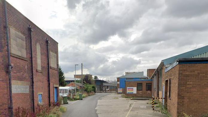 One and two-storey red brick industrial buildings line a partially tarmac-ed street which has weeds sprouting at the sides.