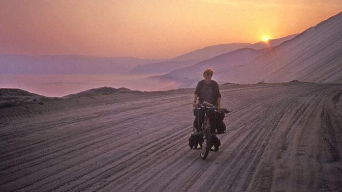 Rona is on her bike. It is loaded with bags. The sandy road is marked by tyre tracks. In the distance the sun is setting behind rugged mountains.