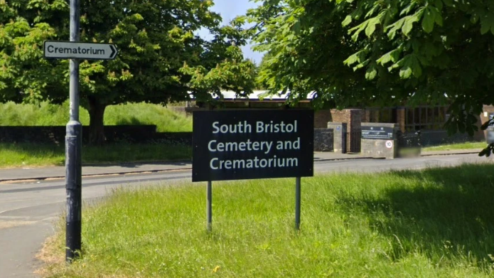 Image shows a sign for South Bristol Cemetery and Crematorium on grass beside a road. 