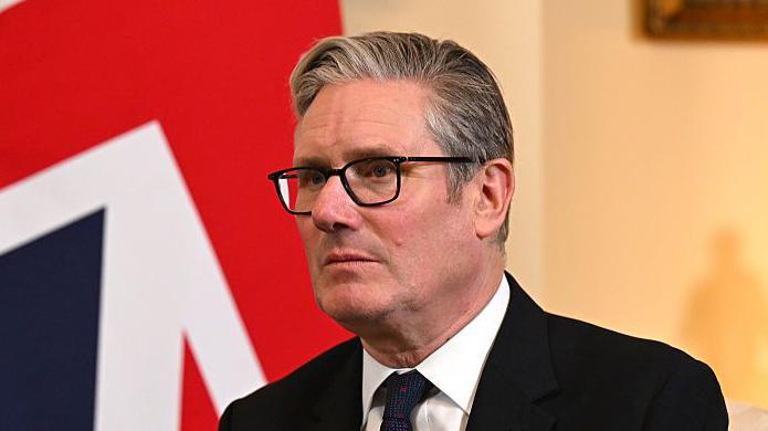 UK Prime Minister Keir Starmer pictured in front of the Union Jack at 10 Downing Street. He wears a dark suit, dark tie and his customary black-framed glasses.
