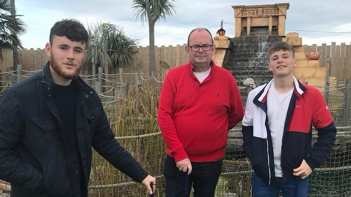 Luke Abrahams with a brown beard, brown hair in a navy coat stands on a mini golf course next to a man with glasses and a red buttoned jumper, next to another man in a red, white and navy coat. 