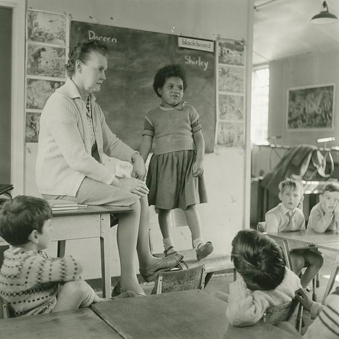 An classroom with four students sat as a desk looking at a teacher, who is sat on a desk with another student standing on a chair next to her.