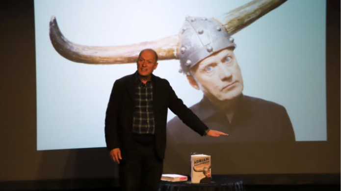 Adrian Edmondson speaking on stage at the Yeovil Literary Festival, with a large picture behind him on a screen of him wear a hat with large horns