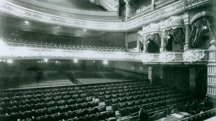 A black and white image of the view from the stage of the theatre, rows of wooden seats make up the stalls, and more can be seen in the circle too. There are individual boxes close to the stage on the circle-level. Ornate designs can be seen on the outside, which connect to front of the balconies.