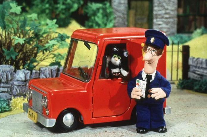 A photograph of Postman Pat taken from the children's programme. Pat has curly brown hair and glasses and is wearing a navy uniform and hat. He is holding a letter in his right hand and standing next to his red delivery van. Jess, his black-and-white cat, is leaning out of the window.