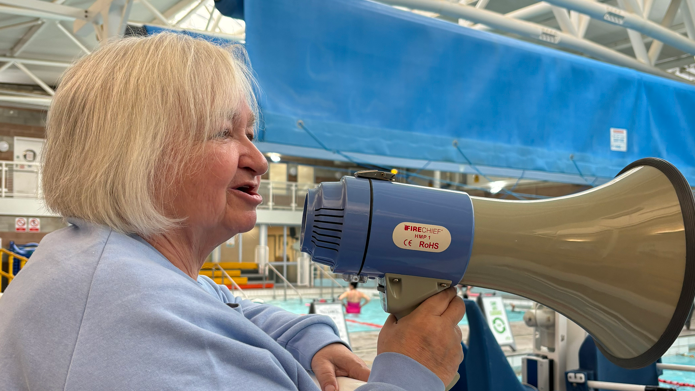 Gemma's mum Sheila Hodgson is stood pool side holding a blue and white megaphone while shouting at Gemma. Sheila has white hair in a bob style haircut, a light blue long sleeve top, and is stood side on to the camera. 