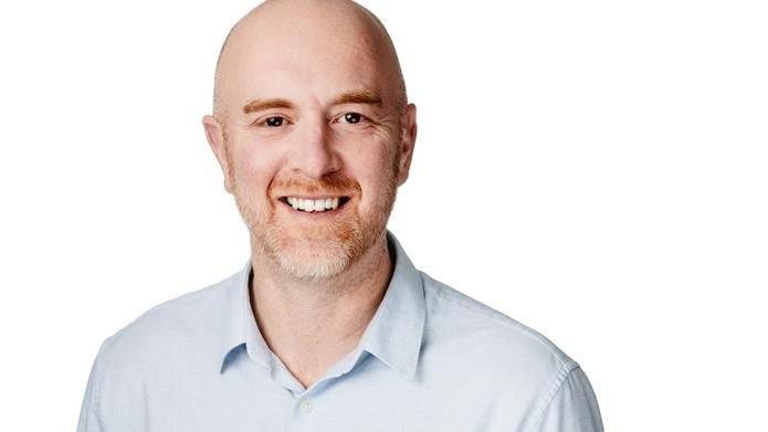 David Powles is wearing a light blue shirt and is smiling. He stands against a white background.