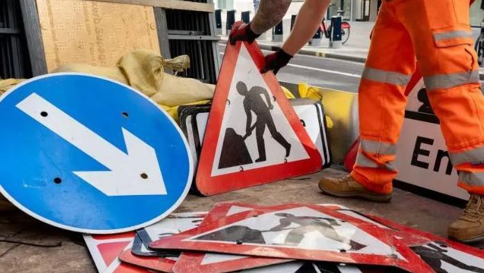 An unidentified road worker wearing hi vis with a roadwork sign and various other roadwork signs around him