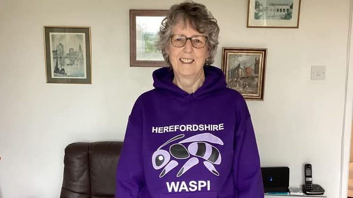 An elderly woman with short grey hair and glasses stands in front of a wall of photos. She is wearing a purple hoodie with a picture of a wasp on the front, which reads "HEREFORDSHIRE WASPI"