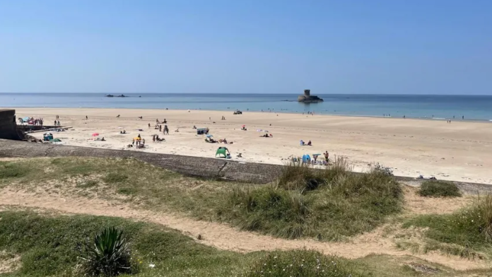 A beach in jersey, in the foreground there are banks of grass situated on a wall overlooking a long sandy beach. Several people have set up on the beach to swim and sunbathe. The sea and horizon can be seen in the distance, the sea is calm with a small rocky outcrop in the middle of the water.