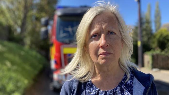 Karen Witton is standing in the foreground in the road and a fire engine is behind her out of focus. She has shoulder length blonde hair, and is wearing a blue and pink top, and a navy blue zip up hoodie.
