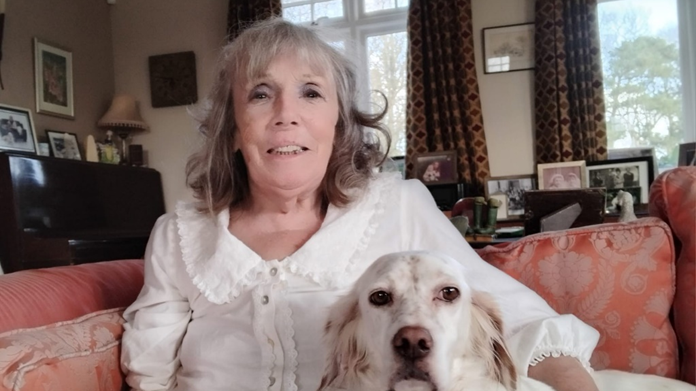 Geraldine at home with her white dog Alice. They are sitting together on a couch. Geraldine is wearing a white blouse and has shoulder length grey hair that is curly at the ends, she is smiling at the camera. Behind her are lots of framed family photos.