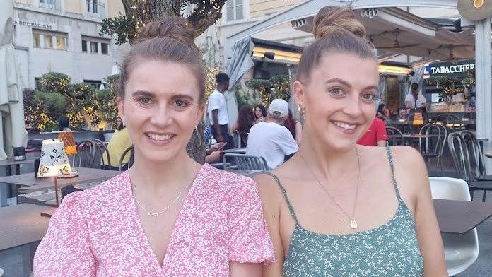Two young women sit at a table in a restaurant, one wearing a pink dress, the other wearing a green dress. Both are looking at the camera.