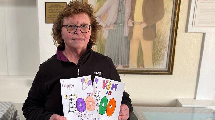 Lindsey Jones holding up hand-drawn cards saying "Happy Birthday Kim" with a picture of a lighthouse drawn on. Lindsey has brown, wavy hair and wears glasses and a dark fleece.