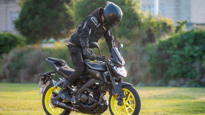 Alfie on a black motorbike with yellow wheels. He's standing out of the saddle wearing all black riding gear and a helmet. The background is a grassy field and hedge.