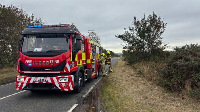 Two firefighters stand next to a fire engine that is parked on a rural road.