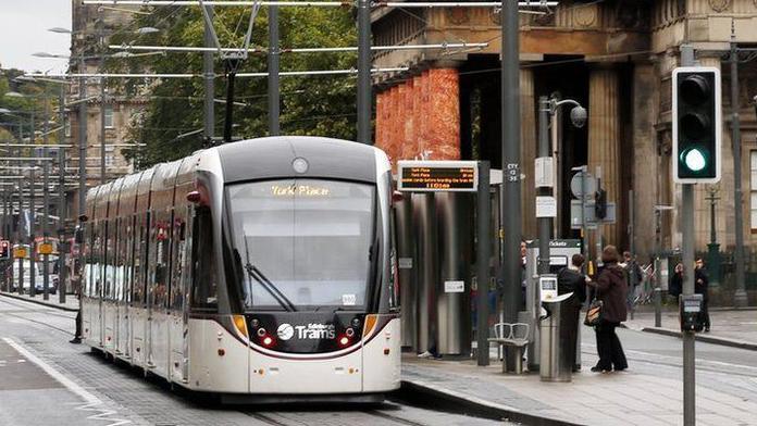 An Edinburgh tram sits at a stop on Princes Street in the capital. In the background is the clock of the Balmoral Hotel, Scott Monument and Calton Hill.