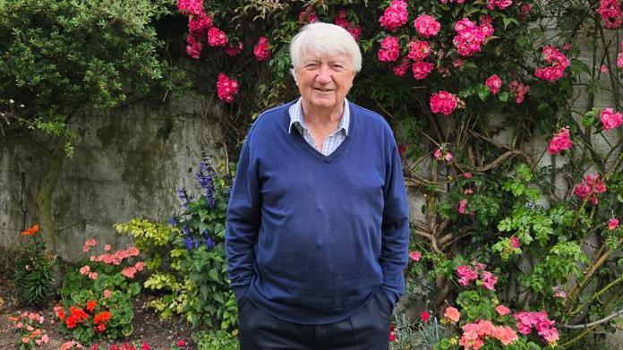 Mid shot of Derrick Downs wearing a blue jumper and smiling to camera standing in front of flowers in a garden 