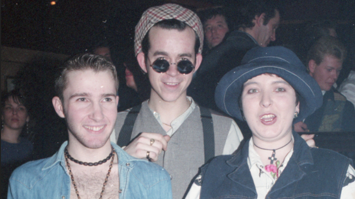 Three people look at the camera. The person on the left wears a light denim, open-neck shirt and necklace. The person in the middle is wearing a hat, sunglasses and grey top. The person on the right is wearing a sleeveless denim jacket and matching hat.