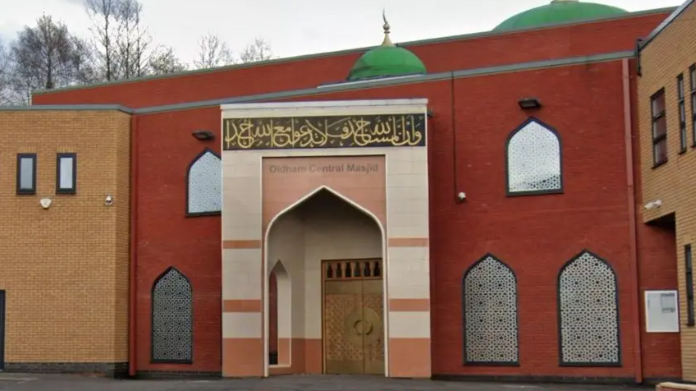 An exterior shot of Oldham Central Mosque- the building - part red brick and part beige brick- has mosaic windows and green domes. Oldhanm Central Majhid is written on the door