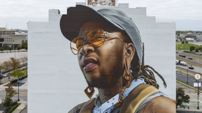 A mural has been painted on the side of a building. It is of a person wearing a grey baseball cap and orange sunglasses.