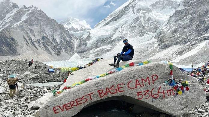 Hari sits with his prostectic legs on a large rock which has the words Everest Base Camp sprayed onto it. Behind him are the mountains and some people who are starting their treks. There are colour ribbons on the rock as well.