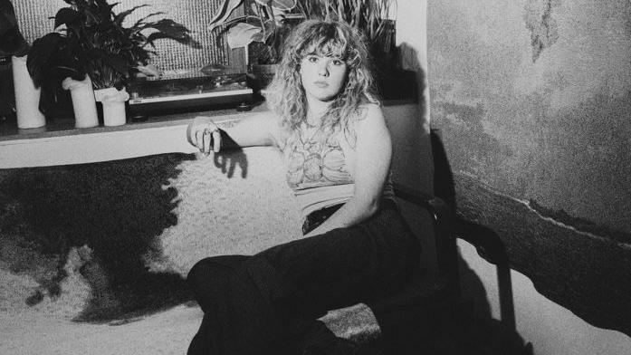 A black and white photo of a young woman with curly long hair and a full fringe sat on a sofa and looking into the camera with a serious expression. Her arm is resting on the side of the sofa and you can see plant pots on a shelf behind her. She is wearing a light vest top and dark trousers.