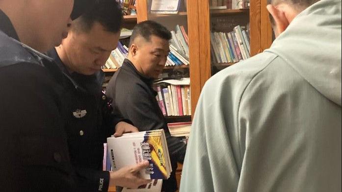 Police officers thumb through books as Sun Cong dressed in black looks on