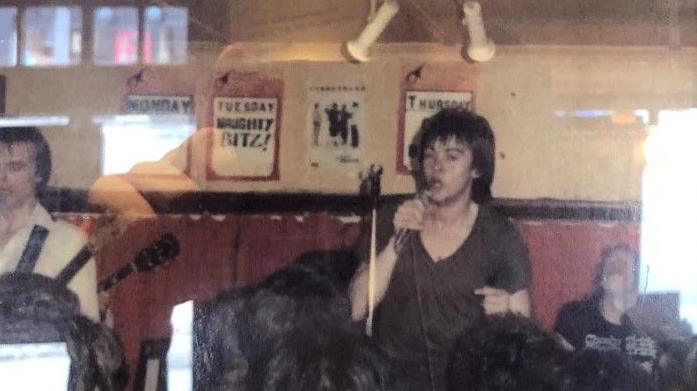 Paul Young in 1979, holding a microphone and singing. The image is old and slightly blurred. A man is sitting down to the right and another man is standing up to the left, playing a guitar. It is in a music venue with posters behind and a red wall halfway up. Peoples' heads are in front of Paul, listening to him perform. 