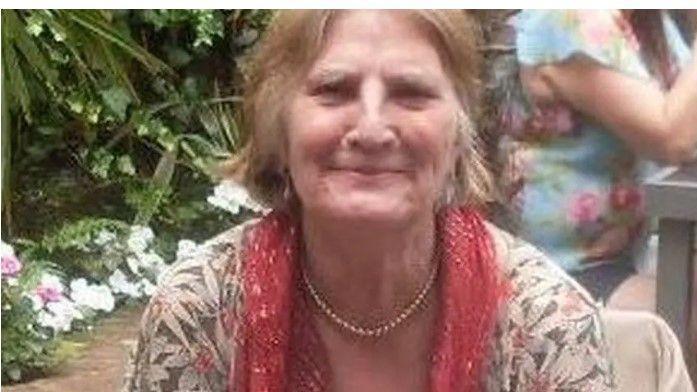 Patricia Holland sits in a garden and is wearing a leaf-patterned jacket, a red scarf and pearl necklace. She is smiling.
