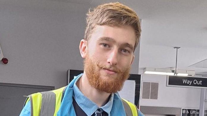 Andrew Mills looking into the camera. He has blonde hair, a beard and a moustache. He has a blue shire on and a yellow hi-viz jacket.