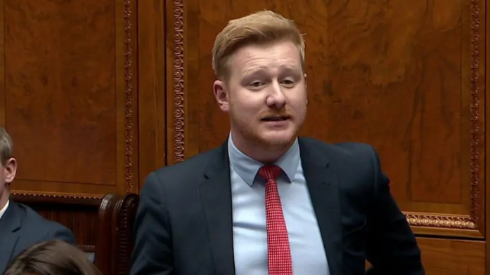 Shows a man with red hair and beard, a blue shirt, red tie, and blue jacket speaking in a debating chamber