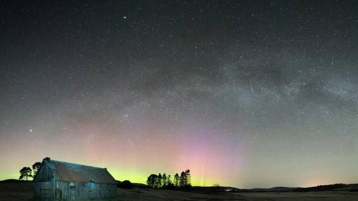 The image shows the Milky Way and Aurora in the sky above Carrbridge along with the trails of four satellites.