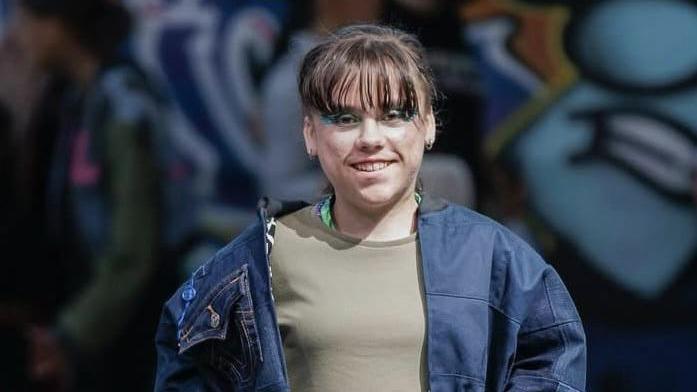 A smiling Carmela Chillery-Watson wearing a grey sweatshirt and an open blue denim jacket. She has long brown hair tied back with a fringe.
